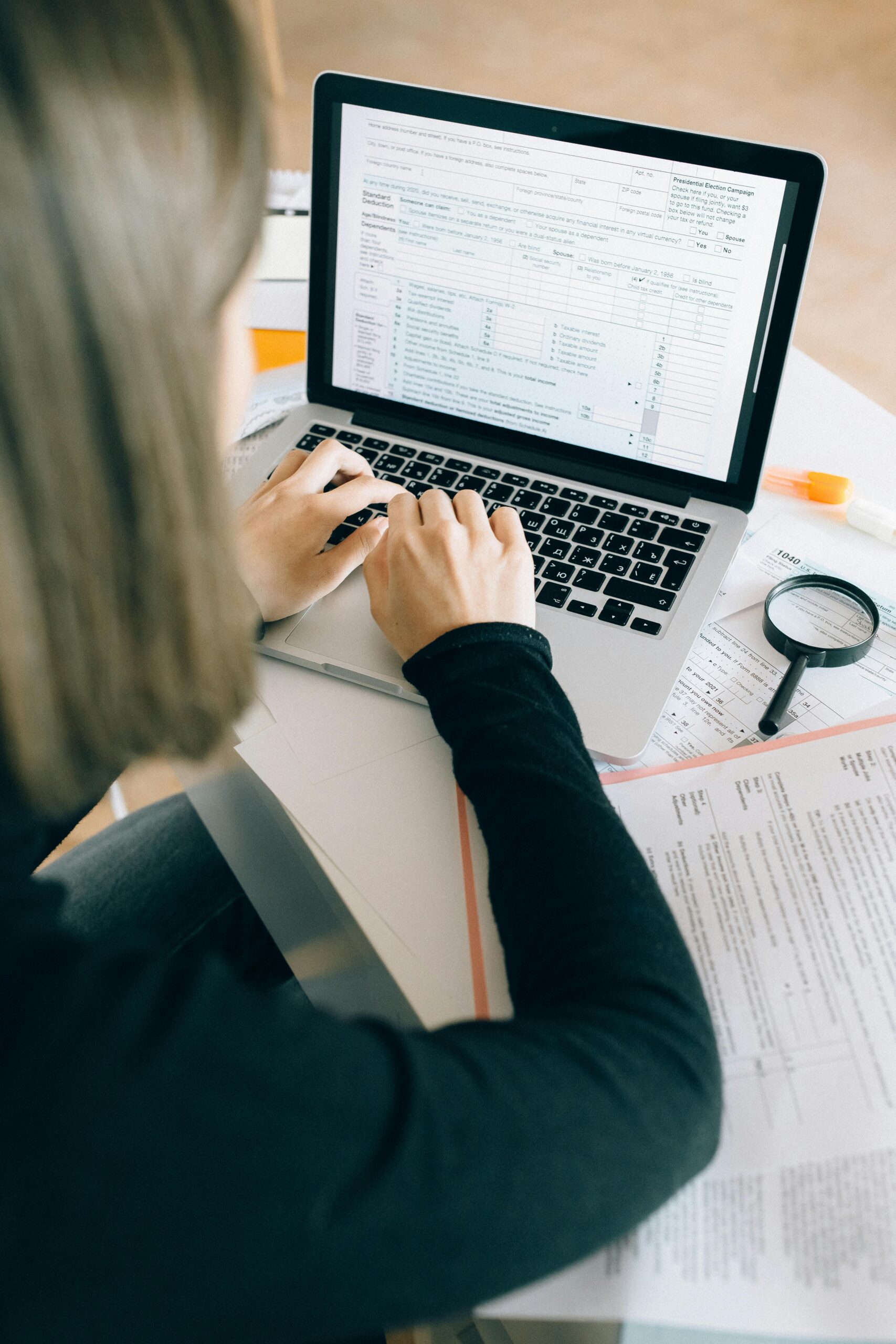 Focused woman working on laptop in office, surrounded by paperwork and magnifying glass.